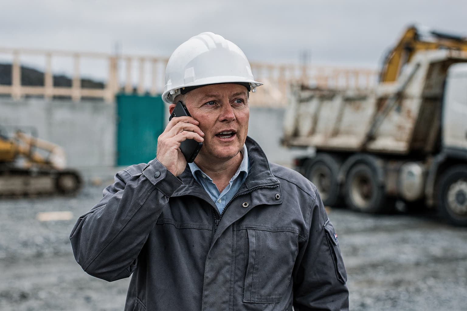 Site manager on the phone on a construction site, with an excavator behind
